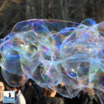 Primo piano di una bolla di sapone gigante composta da più bolle giganti collegate tra loro, con riflessi iridescenti.